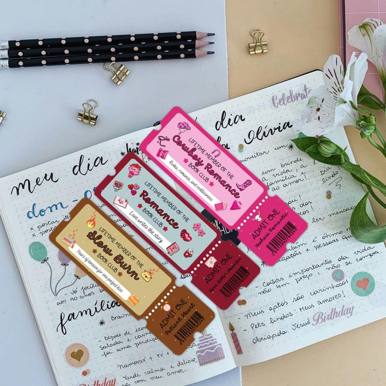 May include: A collection of colorful book club membership cards and admission tickets are displayed on an open journal. The cards feature text like "Cowboy Romance" and "Slow Burn." Pencils and paper clips are also visible.