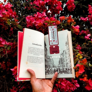 May include: An open book with the title "Introducci&oacute;n" in Spanish, held in front of a backdrop of vibrant red flowers. A bookmark with the text "Romance" and "LITTLE WONDERS THE" is placed inside the book.