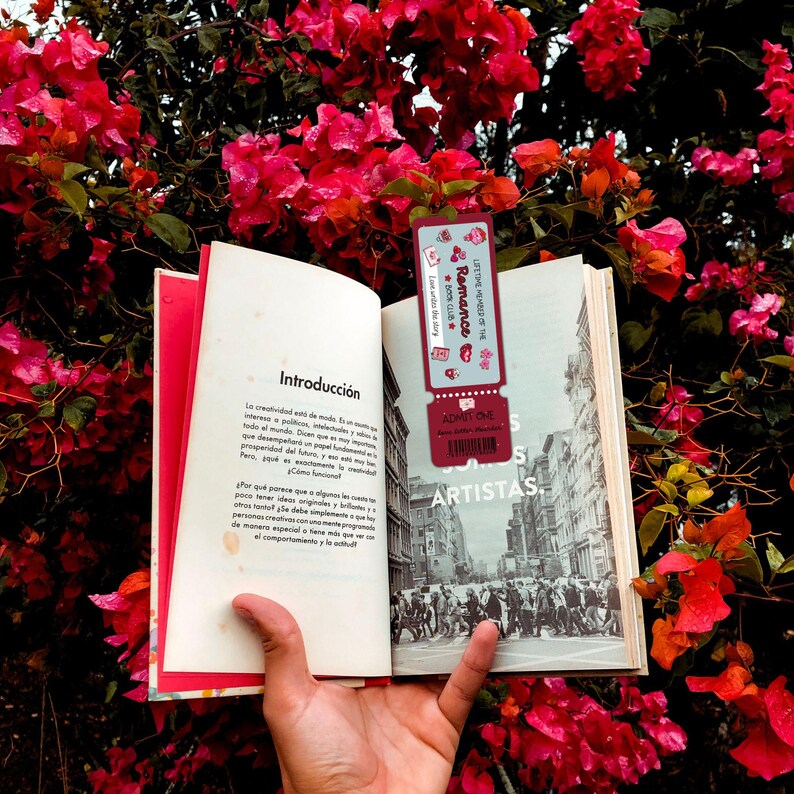 May include: An open book with the title "Introducci&oacute;n" in Spanish, held in front of a backdrop of vibrant red flowers. A bookmark with the text "Romance" and "LITTLE WONDERS THE" is placed inside the book.