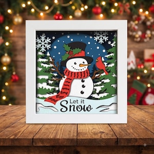 May include: A white-framed shadow box featuring a layered paper cutout of a snowman wearing a black top hat with holly and a red scarf. A red cardinal sits on a pine branch next to the snowman. The text "Let it Snow" is printed at the bottom.
