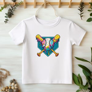 May include: White t-shirt with a graphic of a baseball, two bats, and a home plate. The baseball, bats, and home plate are covered in colorful sprinkles and dripping with pink, blue, and yellow icing.