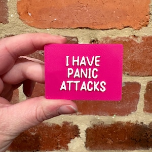 May include: A rectangular hot pink sign with white raised lettering that reads I HAVE PANIC ATTACKS. The sign has a textured surface and is held against a background of red bricks. The text is centred in three lines with a bold sans-serif font.