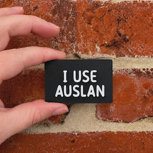 May include: A rectangular black patch with a textured surface featuring the white text I USE AUSLAN in a bold, sans-serif font. The patch has a clean, minimalist design with sharp corners and a matte finish, clearly displaying the message in white.