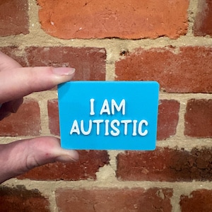Könnte beinhalten: Ein rechteckiges blaues Schild mit weißer, erhabener Schrift, die I AM AUTISTIC lautet. Das Schild hat eine glatte, flache Oberfläche und saubere Kanten, wobei der Text in einer klaren, serifenlosen Schrift auf dem blauen Hintergrund zentriert ist.