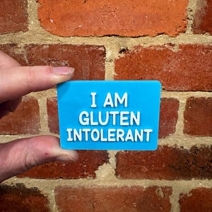 May include: A rectangular blue sign with white raised lettering that reads I AM GLUTEN INTOLERANT. The sign has a smooth, flat surface and features bold, sans-serif text centered on the blue background, creating a clear and legible message.