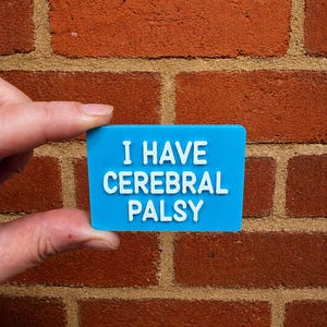 May include: A rectangular blue pin with raised white lettering that reads I HAVE CEREBRAL PALSY. The pin has a smooth, flat surface and a clean, bold font, providing a clear and legible message for communication and awareness purposes.