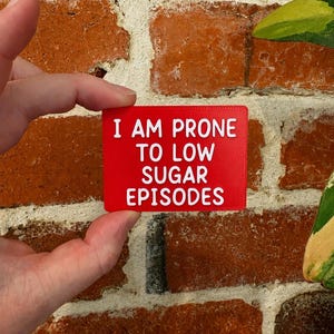 Peut inclure: Un panneau rectangulaire rouge avec des lettres blanches en relief indiquant I AM PRONE TO LOW SUGAR EPISODES. Le panneau présente une surface texturée et est présenté devant un mur de briques rouges et des feuilles vertes, avec un texte clair.