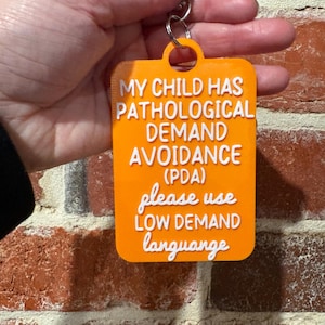 May include: A rectangular orange keychain with rounded corners featuring raised white text that reads: MY CHILD HAS PATHOLOGICAL DEMAND AVOIDANCE (PDA) please use LOW DEMAND language. The tag includes a circular loop at the top with a metal ring.