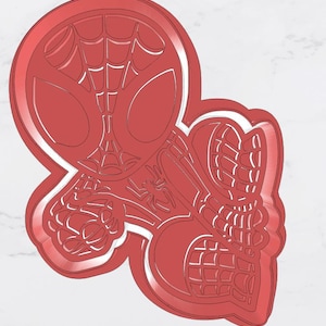 May include: Red cookie cutter in the shape of Spider-Man's head and webbed hands. The design includes the character's mask, spider web details, and a small spider. The cutter is made of a solid red material.