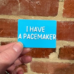 May include: A rectangular bright blue card with white raised lettering that reads I HAVE A PACEMAKER. The card is a flat, solid-colored piece of material with clear, bold text designed for high visibility and easy reading in emergency situations.