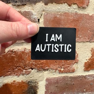 Puede incluir: Un letrero rectangular negro con letras blancas en relieve que dice I AM AUTISTIC. El letrero tiene una superficie texturizada y se sostiene contra un fondo de ladrillos rojos y marrones. El texto está centrado en dos líneas superpuestas.