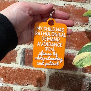 May include: A rectangular orange keychain with white raised text that reads: MY CHILD HAS PATHOLOGICAL DEMAND AVOIDANCE (PDA), please be understanding and patient. The keychain features a circular hole at the top with a metal ring attached.