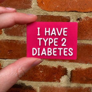 May include: A rectangular hot pink badge featuring the white embossed text I HAVE TYPE 2 DIABETES. The sign has a textured surface and is held in a horizontal orientation, displaying clear lettering in a simple, sans-serif font style.