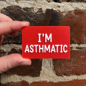 May include: A rectangular red sign with rounded corners featuring the white raised text I'M ASTHMATIC in a bold, sans-serif font. The sign is a flat, solid-colored plaque with a smooth finish and clearly legible white lettering centered on the front.