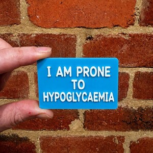 May include: A rectangular blue plastic sign with white raised lettering that reads I AM PRONE TO HYPOGLYCEMIA. The sign features a smooth finish and clear, bold text designed for high visibility and easy reading in a medical context.