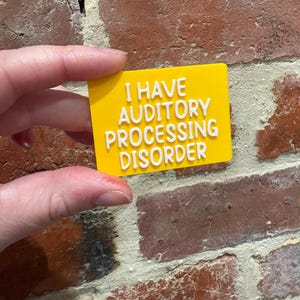 May include: A rectangular yellow sign with raised white lettering that reads I HAVE AUDITORY PROCESSING DISORDER. The sign features a smooth, flat surface with bold, sans-serif text clearly displayed in the center, providing a direct and legible message.
