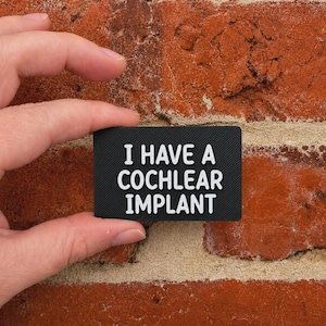 May include: A rectangular black patch with a diagonal ribbed texture featuring the white text I HAVE A COCHLEAR IMPLANT in a bold, sans-serif font. The patch is held against a textured red brick surface, displaying clear, legible lettering.