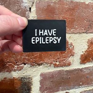 Könnte beinhalten: Ein rechteckiger schwarzer Aufnäher mit dem weißen Text I HAVE EPILEPSY in einer fetten, serifenlosen Schrift. Der Aufnäher hat eine glatte, matte Oberfläche und saubere Kanten, die für eine klare Sichtbarkeit und Identifizierung des Zustands sorgen.