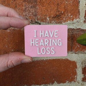 May include: A rectangular pink sign with white raised lettering that reads I HAVE HEARING LOSS. The sign has a smooth, matte finish and is held against a textured red brick wall background, clearly displaying the text for communication.
