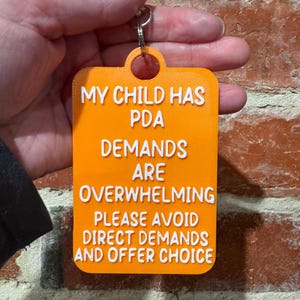 May include: A rectangular orange plastic keychain with rounded corners and a metal ring. The text in raised white lettering reads: MY CHILD HAS PDA DEMANDS ARE OVERWHELMING PLEASE AVOID DIRECT DEMANDS AND OFFER CHOICE. The surface has a textured finish.