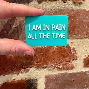 May include: A rectangular teal sign with white raised lettering that reads I AM IN PAIN ALL THE TIME. The sign has a smooth, flat surface and a clean, modern font style, presenting a clear and legible message in a bold, high-contrast colour scheme.