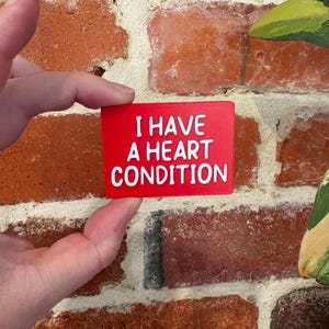 May include: A rectangular red sign with white raised lettering that reads I HAVE A HEART CONDITION. The sign has a textured surface and is held in place against a background of red bricks. The text is clearly legible and centered on the sign.