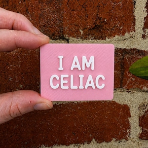 May include: A rectangular pink sign with white raised lettering that reads I AM CELIAC. The sign has a smooth, flat surface and features bold, sans-serif text centered on the front face, providing a clear and legible message for identification.