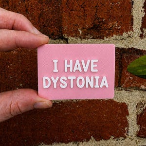 May include: A rectangular pink sign with white raised lettering that reads I HAVE DYSTONIA. The sign has a textured surface and is held against a red brick background. The text is centered in two lines with the words stacked vertically.