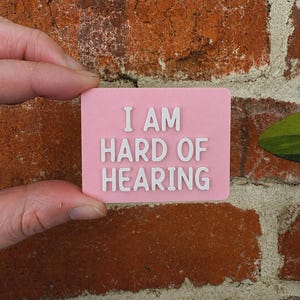 May include: A rectangular pink sign with raised white lettering that reads I AM HARD OF HEARING. The sign has a smooth, matte finish and features clean, bold, sans-serif typography centered on the surface, providing a clear and legible message.