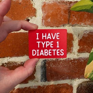 May include: A rectangular red medical alert sign with raised white lettering that reads I HAVE TYPE 1 DIABETES. The sign has a textured surface and is held in a horizontal orientation, featuring clear, bold, sans-serif text for high visibility.