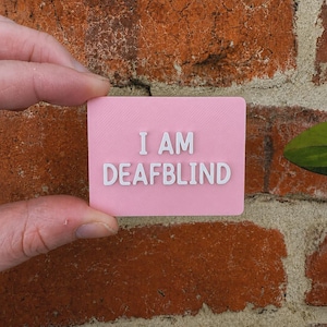 May include: A rectangular pink sign with rounded corners featuring the white raised text I AM DEAFBLIND centered on the front surface. The sign has a matte finish and a textured appearance, displaying clear lettering in a bold, sans-serif font style.