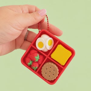 May include: A red, square-shaped keyring with a divided compartment containing miniature food items. The compartments hold strawberries, hard-boiled eggs, a slice of cheese, and a biscuit. The keyring is held by a red chain.