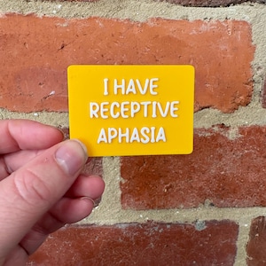 May include: A rectangular yellow sign with raised white lettering that reads I HAVE RECEPTIVE APHASIA. The sign features a smooth, flat surface with bold, clear text designed for communication assistance and accessibility purposes.