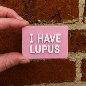 May include: A rectangular pink sign with rounded corners featuring the white raised text I HAVE LUPUS in a bold sans-serif font. The surface of the pink plastic has a fine, linear 3D printed texture visible across the entire background area.