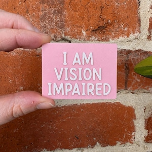 May include: A rectangular pink sign with raised white lettering that reads I AM VISION IMPAIRED. The sign has a smooth, matte finish and features clean, sans-serif typography centered on the surface, providing a clear and legible message.