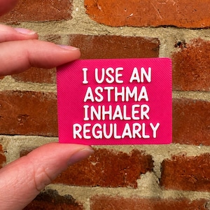 Peut inclure: Un panneau rectangulaire rose vif avec des lettres blanches en relief indiquant I USE AN ASTHMA INHALER REGULARLY. Le panneau présente une surface texturée et est tenu devant un mur de briques rouge-brun avec des joints de mortier visibles.