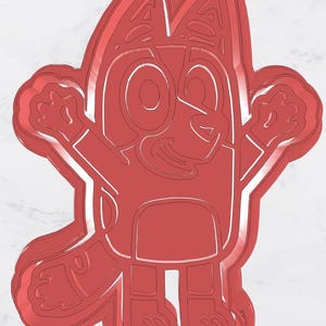 Peut inclure: Un emporte-pièce rouge en forme de chien de dessin animé, avec de grandes oreilles, les bras levés et un visage souriant. Le découpoir a des contours détaillés et est conçu pour la pâtisserie.