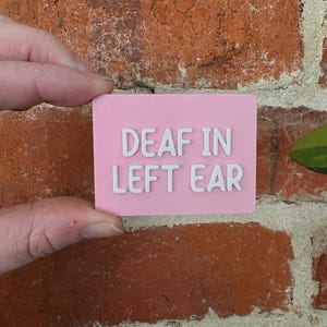 May include: A rectangular pink sign with white raised lettering that reads DEAF IN LEFT EAR. The sign has a smooth, matte finish and features clean, bold sans-serif typography centered on the surface, providing a clear and legible message.