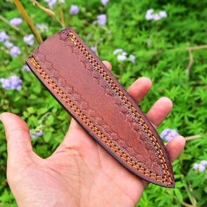 May include: A brown leather knife sheath with decorative stitching. The sheath has a pointed tip and a flap that closes with a snap.
