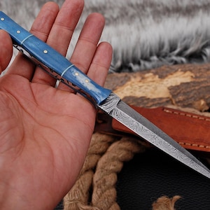 May include: A small, ornate knife with a blue handle and a patterned blade is held in a hand. The handle has silver accents. The blade has a unique, textured design. The text "X - 113" is visible in the upper right corner.