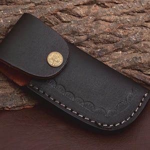 May include: A black leather knife sheath with a decorative scalloped edge and white stitching. The sheath has a flap secured by a gold-colored button. The interior is a lighter brown color.