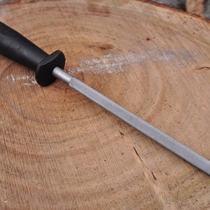 May include: A black handled knife sharpener with a silver sharpening rod. The sharpener has a ring at the end of the handle. The tool is resting on a wooden surface.