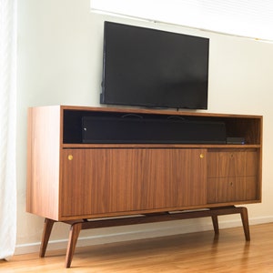 Puede incluir: Mueble de TV de estilo moderno de mediados de siglo en nogal. Tiene un estante superior, una puerta corredera, dos cajones y patas anguladas. Una barra de sonido y un televisor negros est&aacute;n encima. Aproximadamente 150 cm de ancho.