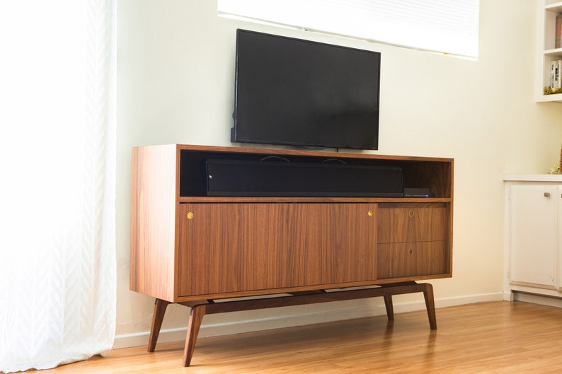 Puede incluir: Mueble de TV de estilo moderno de mediados de siglo en nogal. Tiene un estante superior, una puerta corredera, dos cajones y patas anguladas. Una barra de sonido y un televisor negros est&aacute;n encima. Aproximadamente 150 cm de ancho.