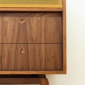 Puede incluir: Un mueble de madera de estilo moderno de mediados de siglo con dos cajones y una secci&oacute;n de altavoz. El mueble es de madera oscura con una cubierta de altavoz de tela amarilla y negra. Los cajones tienen tiradores circulares.