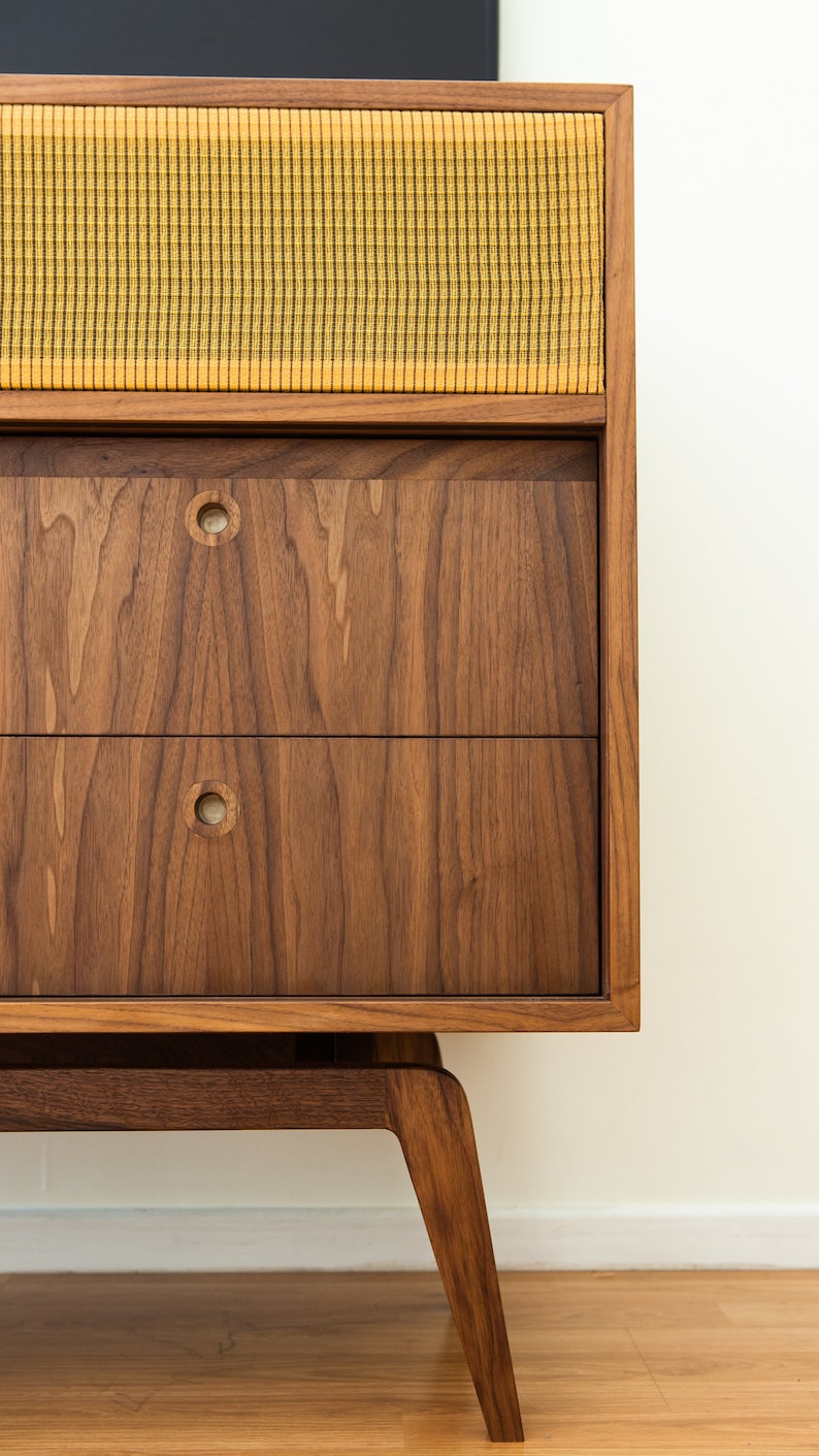 Puede incluir: Un mueble de madera de estilo moderno de mediados de siglo con dos cajones y una secci&oacute;n de altavoz. El mueble es de madera oscura con una cubierta de altavoz de tela amarilla y negra. Los cajones tienen tiradores circulares.