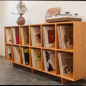 May include: A light wood record shelf with multiple compartments filled with vinyl records. A record player sits on top. A vase with dried flowers is on the top left. The shelf is against a white wall.