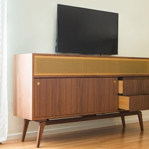 Puede incluir: Mueble de TV de madera de estilo moderno de mediados de siglo con un televisor de pantalla plana negro encima. El mueble presenta un acabado de nogal, detalles de lat&oacute;n y una rejilla de altavoz. Dos cajones est&aacute;n abiertos.
