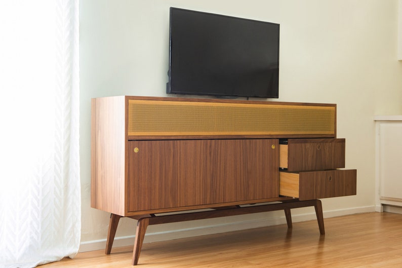 Puede incluir: Mueble de TV de madera de estilo moderno de mediados de siglo con un televisor de pantalla plana negro encima. El mueble presenta un acabado de nogal, detalles de lat&oacute;n y una rejilla de altavoz. Dos cajones est&aacute;n abiertos.