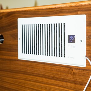 Puede incluir: Una rejilla de ventilaci&oacute;n AC Infinity blanca con lamas verticales y una pantalla digital que muestra 30&deg;C. La rejilla est&aacute; montada sobre una superficie de madera oscura. Un cable blanco sale de la rejilla.